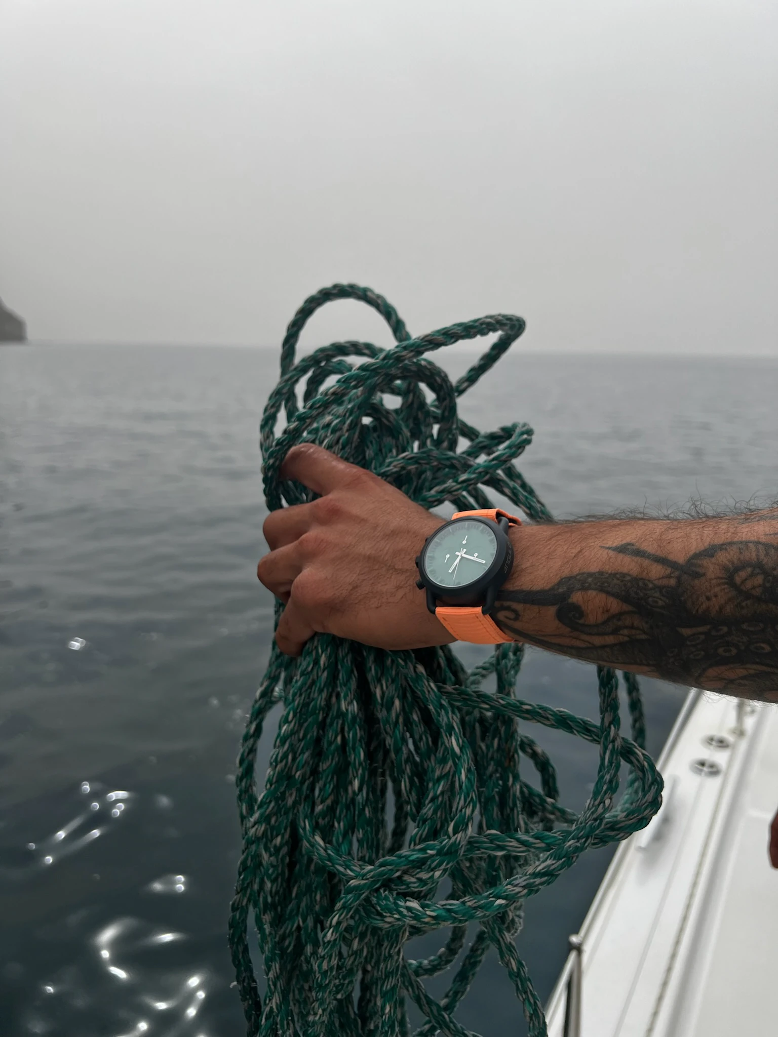 Hand med en Angulo-klocka som håller en rulle rep över havet från en segelbåt